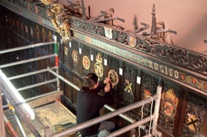 A conservator works from a scaffolding tower on a series of colourful heraldic designs on timber panelling