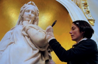 A conservator brushes microcrstalline wax onto a marble statue in an alcove lined with gold leaf or gold paint