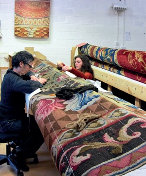 Two female conservators in a workshop repair a historic carpet mounted on special rollers