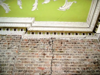 Interior wall with plaster removed exposing brick with long vertical crack disappearing at cornice