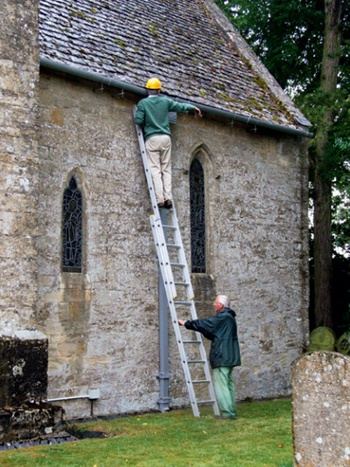 Man on a ladder and wearing a hard-hat inspects a church gutter