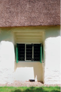 The crisp edge of a thatched church roof overhanging a recessed window with green exterior shutters