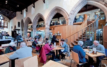 A busy cafeteria in a historic church nave with a modern staircase in the background leading to a first floor gallery