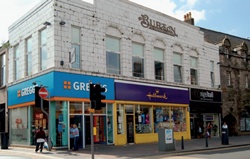 Former Burton’s store retaining Burton's logo above upper storey windows; ground floor shops (Greggs, Hallmark, etc) have brightly coloured modern fronts